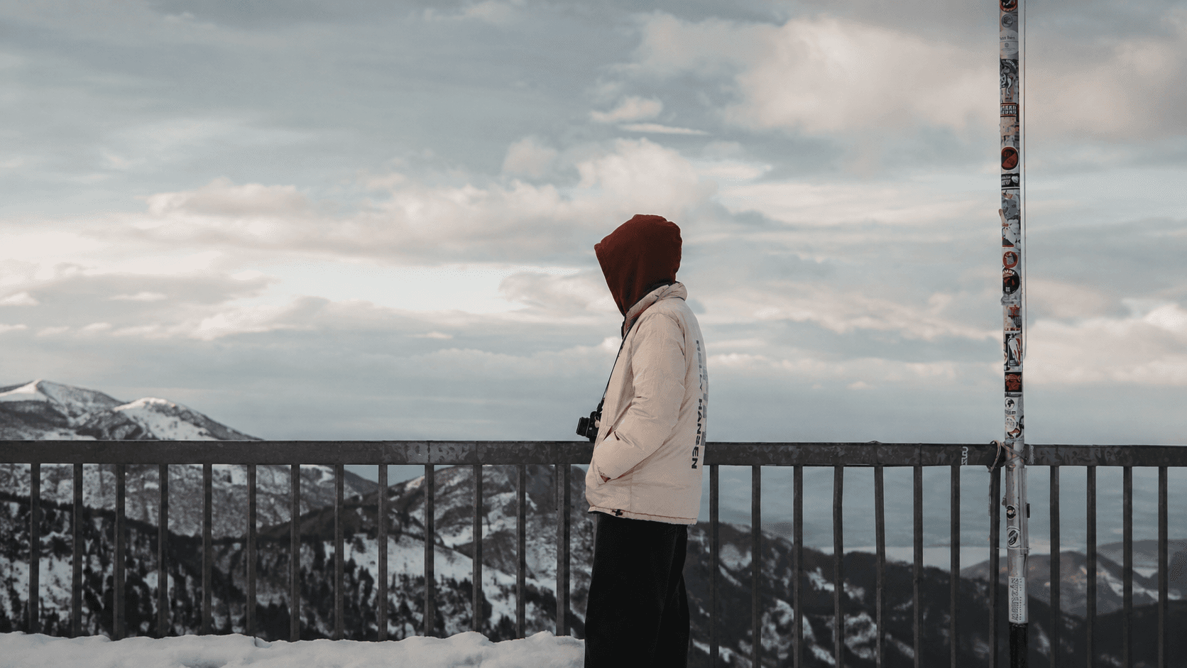 A hooded figure in a cream jacket stands at a railing above snowy mountains under a cloudy sky, camera in hand.